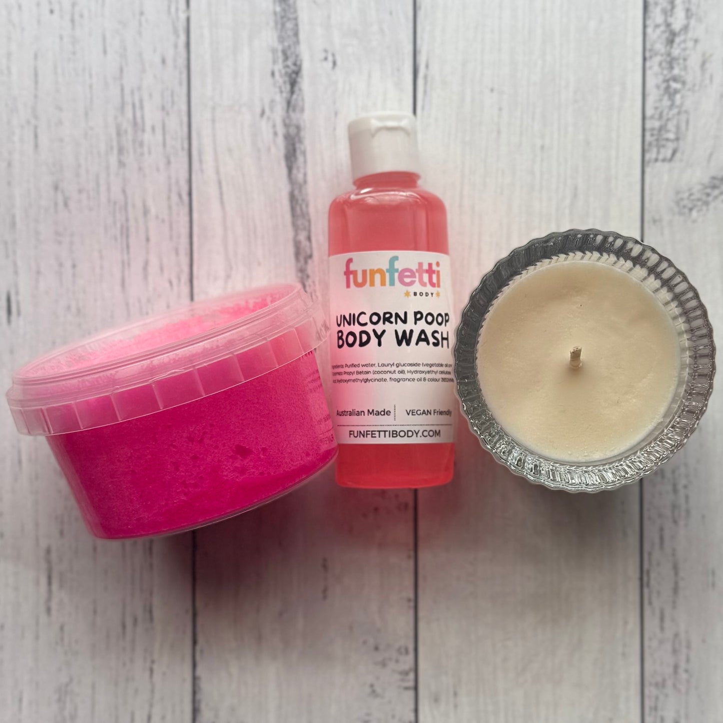 Pink container, bottle of 'funfetti' unicorn poop body wash, and candle on a wooden surface.
